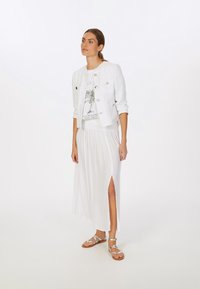 Weiße Leinenjacke mit silbernen Knöpfen, kombiniert mit einem weißen Grafik-T-Shirt und einem fließenden weißen Rock mit seitlichem Schlitz, getragen mit silbernen Sandalen.