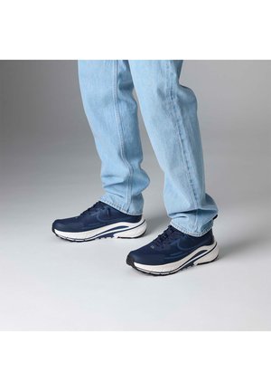 Person, die hellblaue Jeans und dunkelblaue Sportschuhe trägt, steht auf einer hellgrauen Oberfläche.
