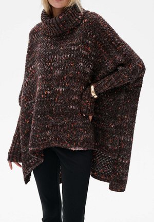 Kobieta wearing a chunky, speckled brown turtleneck sweater z asymetrycznym dołem i długimi rękawami, w połączeniu z czarnymi spodniami i złotą bransoletką.