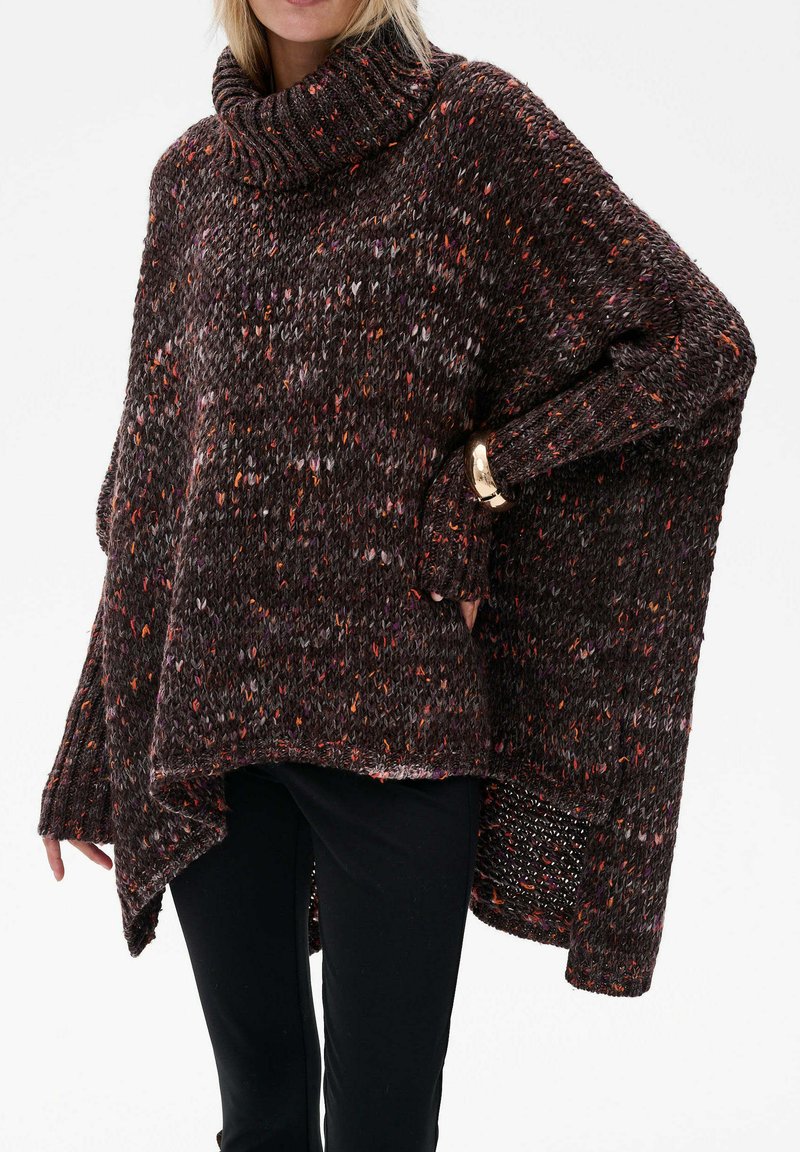 Kobieta wearing a chunky, speckled brown turtleneck sweater z asymetrycznym dołem i długimi rękawami, w połączeniu z czarnymi spodniami i złotą bransoletką.