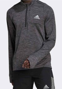 Chemise de sport gris à fermeture éclair partielle, manches longues, tissu texturé, avec une petite poche avant ornée du logo Adidas blanc.