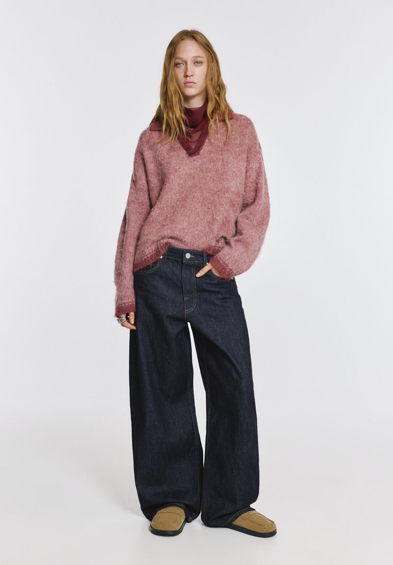 Vastag, rózsaszín kötött pulóver V-nyakkal, széles szárú sötét farmernadrággal párosítva. A modell bézs slip-on cipőt visel. Egyszerű, lezser összeállítás.