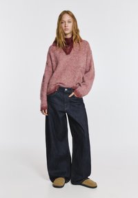 Pulover gros, roz, tricotat, cu decolteu în V, combinat cu blugi largi din denim închis la culoare. Modelul poartă pantofi slip-on de culoare maro deschis. Ansamblu simplu și casual.