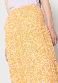 Lähedalt pildista isikut, kes kannab kõrge vöökohtaga kollast seelikut, millel on valge abstraktne muster ja helesinine topp, näidates külge ja kätt.