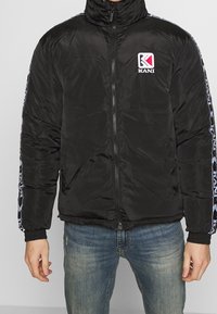 Svart pufferjacka med hög krage, dragkedja och ribbade mudd. Har en logotyp på bröstet och vita mönstrade detaljer på ärmarna.