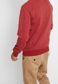 Sudadera texturizada roja con mangas largas y puños de canalé, combinada con pantalones caqui con bolsillos y botones negros.