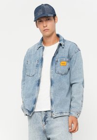 Light blue denim jacket with two front pockets, button closure, and yellow label. Worn over a white t-shirt, paired with matching denim.
