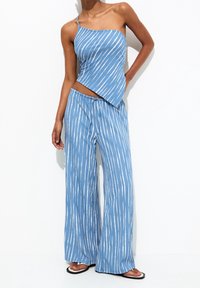 Helles blaues zweiteiliges Outfit mit einem schräg geschnittenen Spaghetti-Träger-Top und lockeren, weit geschnittenen Hosen, die weiße diagonale Streifen aufweisen.