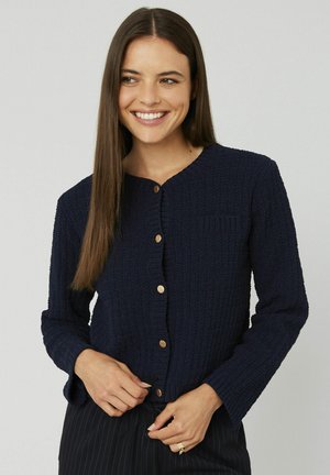 Cardigan texturé bleu marine avec un col rond, des manches longues et une fermeture à boutons en bois, comportant une petite poche poitrine et des poignets côtelés.