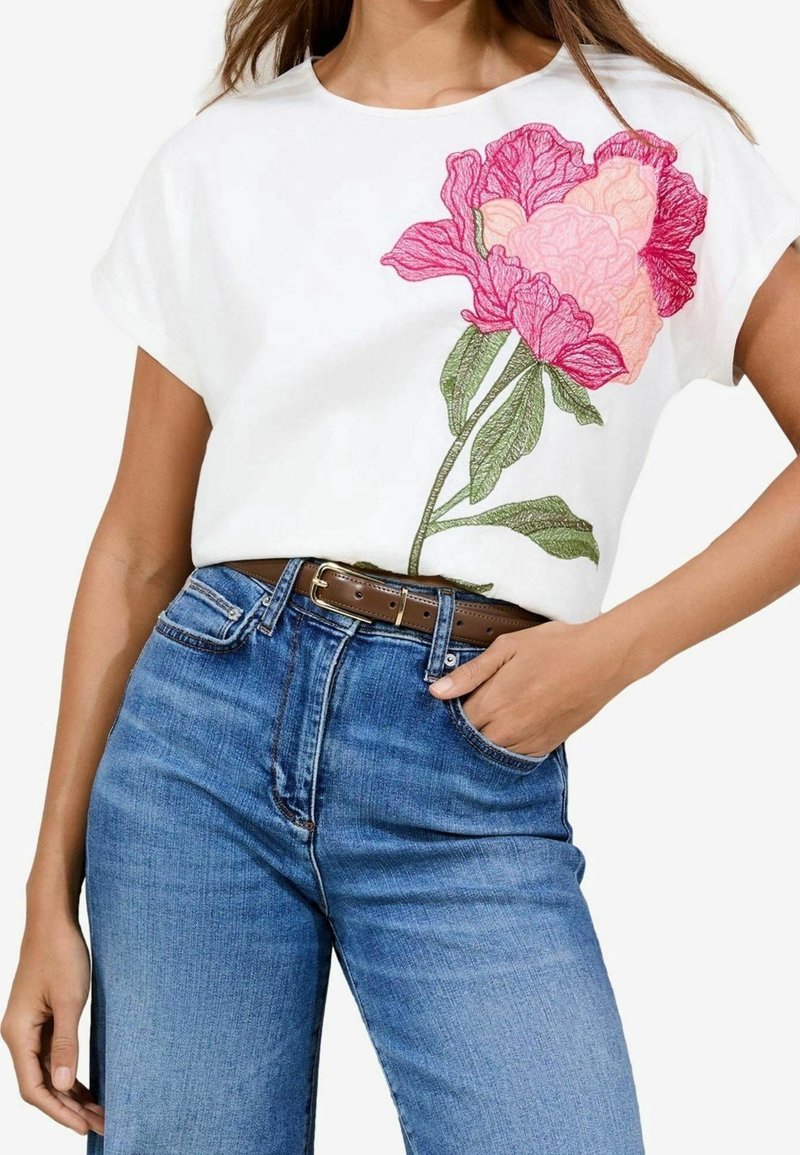 Femme portant un haut blanc à manches courtes avec une grande broderie de fleur rose, associé à un jean bleu taille haute et une ceinture marron.