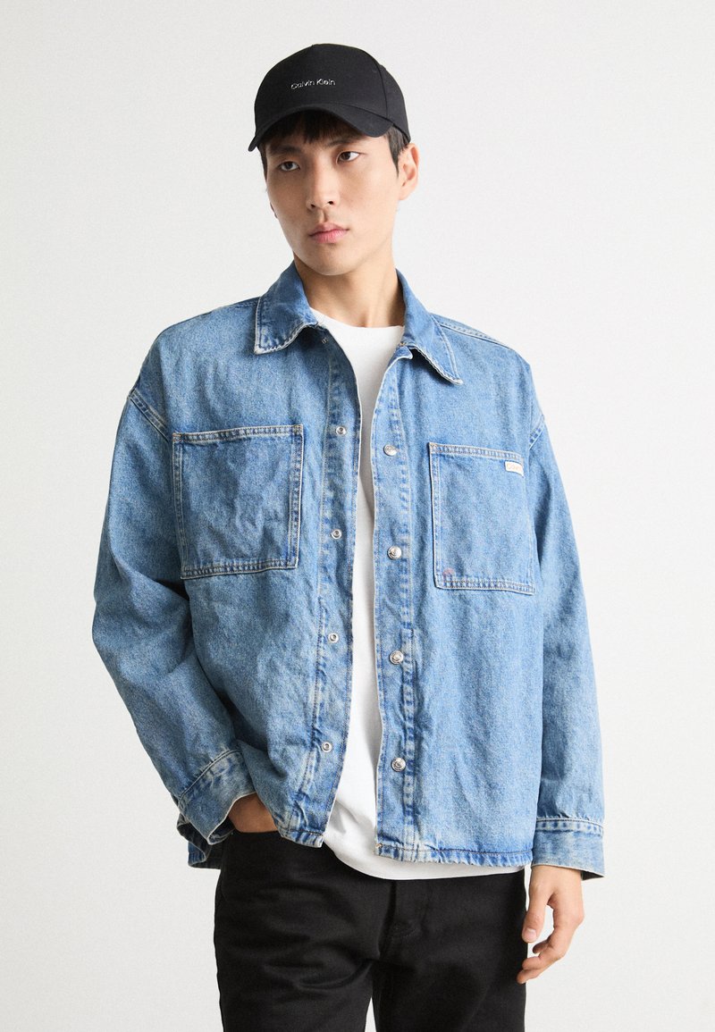 Lichtblauwe denimjas met twee voorzakken, knoopsluitingen en een losse pasvorm, gedragen over een wit shirt en aangevuld met een zwarte pet.