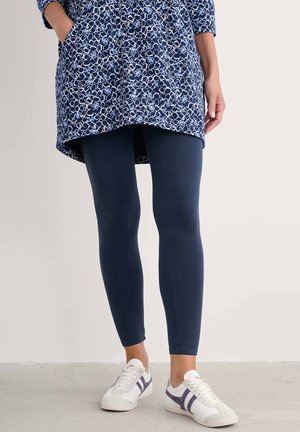 Marineblauwe tuniek met wit abstract patroon, voorzakken, gecombineerd met marineblauwe leggings en witte sneakers met paarse accenten.