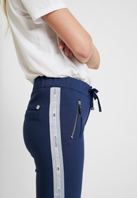 Mörkblå byxor med en texturerad tyg, med dragsko i midjan, sidofickor med dragkedja och en ljusblå accentrand med logodetalj.