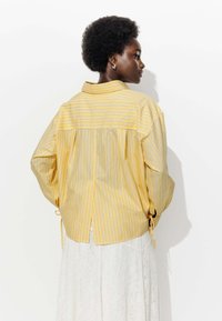 Chemise rayée jaune et bleue à coupe décontractée, à manches longues et avec un pli au dos. Présente des nœuds sur les côtés et un col classique.