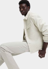 Jasnobeżowa wełniana kurtka o stonowanym kroju, z dwoma przednimi kieszeniami i ściągaczem na mankietach, noszona wraz z dopasowanymi jasnymi spodniami. Prosta i nowoczesna.