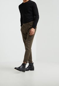 Schwarzes Langarmshirt, olivgrüne maßgeschneiderte Hose und schwarze Knöchelstiefel mit strukturierten Seiten. Die Hose ist an den Knöcheln umgeschlagen.