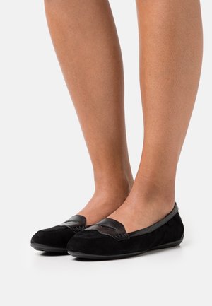 Paire de mocassins en daim noir avec garniture en cuir texturé portés pieds nus, présentés sur un fond blanc.