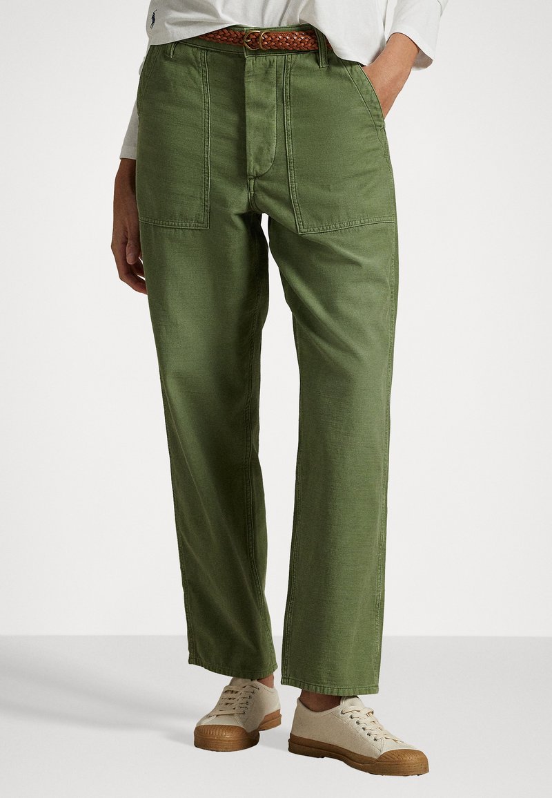 Pantaloni di cotone verdi con taglio dritto, dotati di due grandi tasche anteriori e una cintura intrecciata marrone. Indossati con sneakers color beige chiaro.
