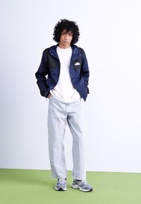 Jūras spēku vējjakas ar melniem akcentiem, balta T-kreklu, gaiši zili džinsi un pelēki sporta apavi, pozējot uz neitrāla fona.