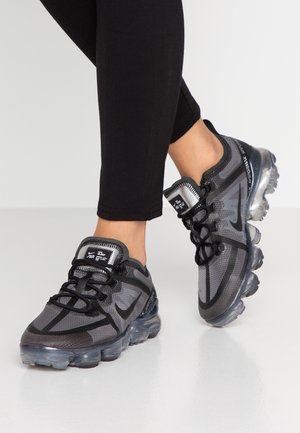 Person iført sorte leggings og mørkegrå Nike Air VaporMax sneakers med gennemsigtige boblesåler, stående på et lyst gulv.