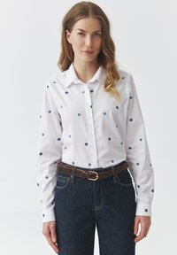 Witte button-up shirt met blauwe geborduurde bloemmotieven, lange mouwen en een klassieke kraag, gecombineerd met donkerdenim jeans en een leren bruine riem.