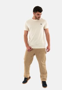 T-shirt beige a maniche corte con scollatura rotonda e logo ricamato, abbinato a pantaloni cargo color sabbia con molteplici tasche e sneakers nere.
