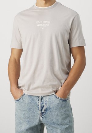 Homme portant un t-shirt beige clair Emporio Armani Milano légèrement rentré dans un jean en denim bleu clair, les mains dans les poches.