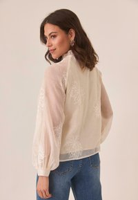 Blusa blanca bordada con mangas largas y abullonadas. Tejido transparente, patrones florales, cuello alto. Combinada con pantalones vaqueros azules.