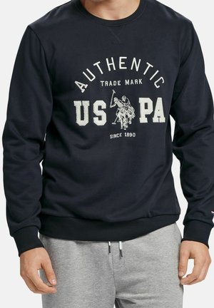 Mężczyzna ubrany w czarną bluzę z białym napisem "AUTHENTIC US PA" i grafiką gracza polo, w połączeniu z jasnoszarymi spodniami dresowymi.