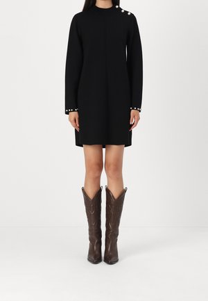 Personne portant une robe noire à manches longues avec des boutons en perle sur l'épaule et les poignets, associée à des bottes en cuir marron montant jusqu'au genou.