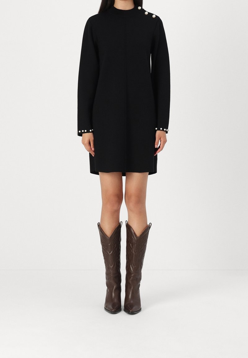 Personne portant une robe noire à manches longues avec des boutons en perle sur l'épaule et les poignets, associée à des bottes en cuir marron montant jusqu'au genou.