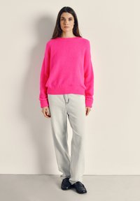 Donna in piedi contro un muro beige che indossa un maglione rosa acceso, jeans chiari e mocassini neri, rivolta verso l'obiettivo con un'espressione neutra.