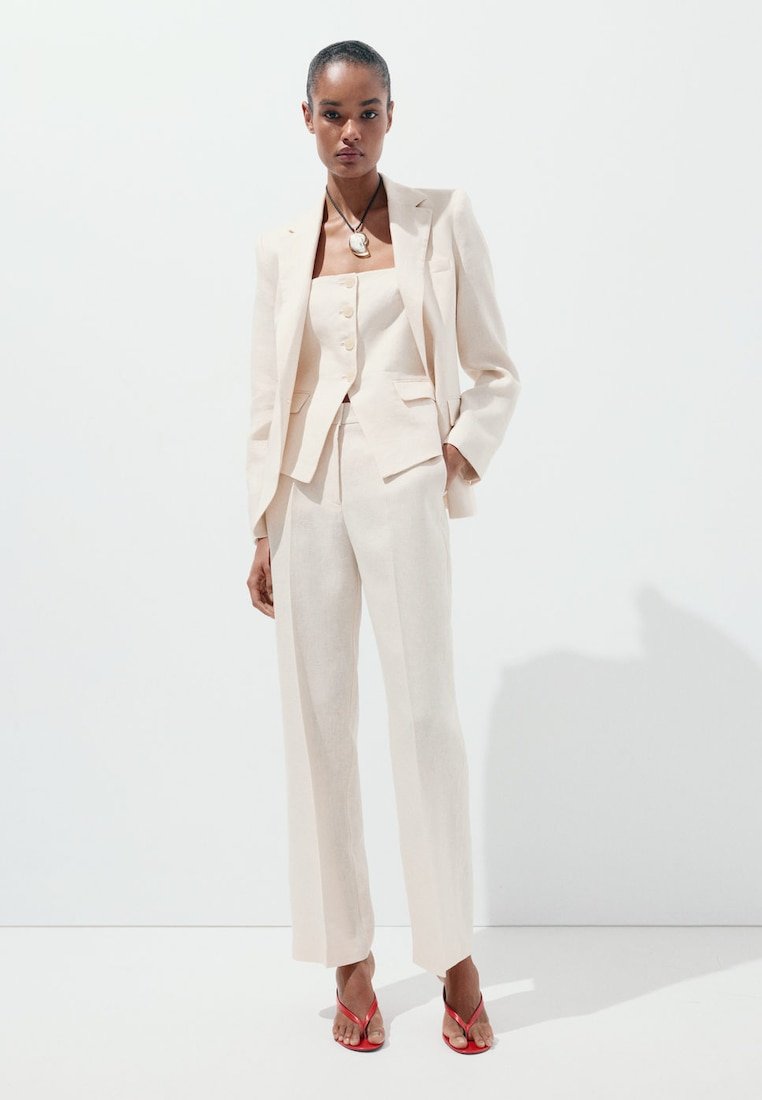 Femme debout portant un costume trois pièces couleur crème avec veste, gilet boutonné, pantalon à jambes larges, talons rouges ouverts et collier pendentif.