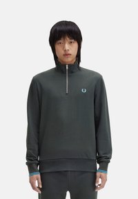 Sudadera de cuello alto de color verde oscuro con una textura suave, que presenta un pequeño logo azul y acentos azules contrastantes en los puños.