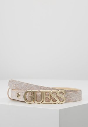 Schmaler beiger Gürtel mit Muster und großer goldener "GUESS"-Schließe, der auf einer weißen Oberfläche vor einem schlichten grauen Hintergrund liegt.