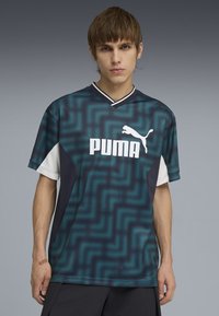 Giovane uomo che indossa una maglietta Puma blu e nera con motivo a fantasia e logo bianco, in piedi davanti a uno sfondo sfumato grigio.