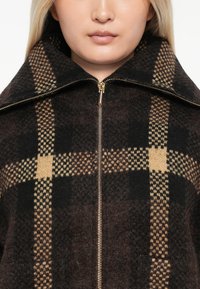 Braun- und schwarz karierter Reißverschlussjacke mit hohem Kragen und goldenen Hardware-Elementen. Weicher, strukturiert Stoff mit großen quadratischen Mustern.