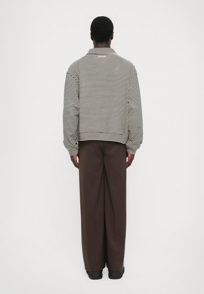 Chemise à manches longues rayée en noir et blanc, avec un col, portée avec un pantalon large marron. Texture lisse, coupe décontractée, détails minimalistes.