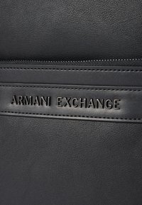 Sac en cuir noir texturé avec un logo "ARMANI EXCHANGE" en relief. Comprend un détail de fermeture éclair et des accents de couture subtils.