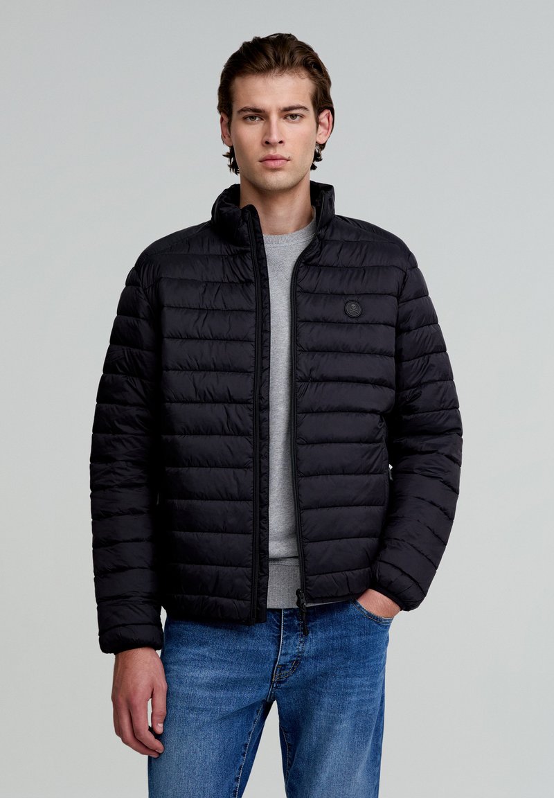 Sort pufferjakke med quiltet tekstur, høj krave og lynlås foran. Parret med en grå sweatshirts og blå jeans.