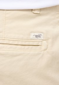 Pantalons en coton beige avec une poche arrière arborant un patch logo gris. Le tissu est lisse avec une texture subtile et des détails de couture soignés.