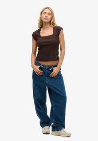 Top corto marrón con bordes en forma de ola, combinado con jeans de mezclilla azul de corte holgado y zapatillas de color claro. Diseño sencillo, ajuste casual.