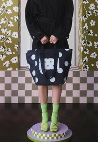 Denimu soma ar lieliem baltiem punktiņiem un zilu kabatu. Iekļauj tumšas rokturus, sarkanu logo un atrodas uz apļa krāsota pjedestāla. Redzami zaļi zeķes.