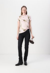 T-shirt floral rosa com mangas curtas, combinada com jeans pretos e botas de tornozelo pretas. Modelo segura uma pequena mala castanha.