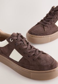 Baskets en daim marron avec des panneaux latéraux blancs, des semelles épaisses en caoutchouc beige et des lacets marron sur un fond clair.