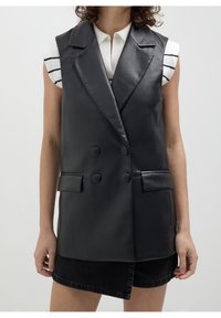 Svart faux läder ärmlös blazer med dubbelknäppta knappar, skarpa slag och fickor, bärs över en randig ärmtröja och denimshorts.