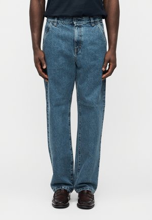 Man wearing loose-fit blue denim jeans and dark brown leather loafers, standing against a plain white background.