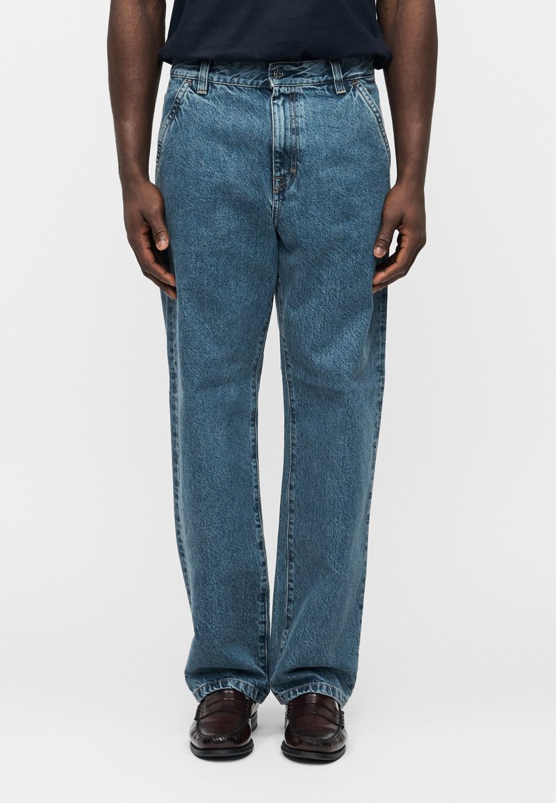 Άνδρας με φαρδιά μπλε τζιν και σκούρα καφέ δερμάτινα μοκασίνια, που στέκεται μπροστά σε απλό λευκό φόντο.