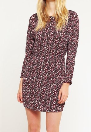 Kobieta ubrana w sukienkę z długim rękawem sięgającą kolan, zdobioną małym kwiatowym wzorem w kolorach czerwonym, białym i czarnym na ciemnym tle.