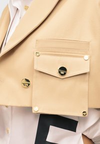 Μπεζ σακάκι με μεγάλη τσέπη με πτερύγιο, κλειστή με στρογγυλά χρυσά κουμπιά, πάνω από λευκό πουκάμισο με μαύρη γραφική λεπτομέρεια.